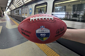 Fotografia de uma pessoa segurando uma bola de futebol americano, dentro da estação de metrô.